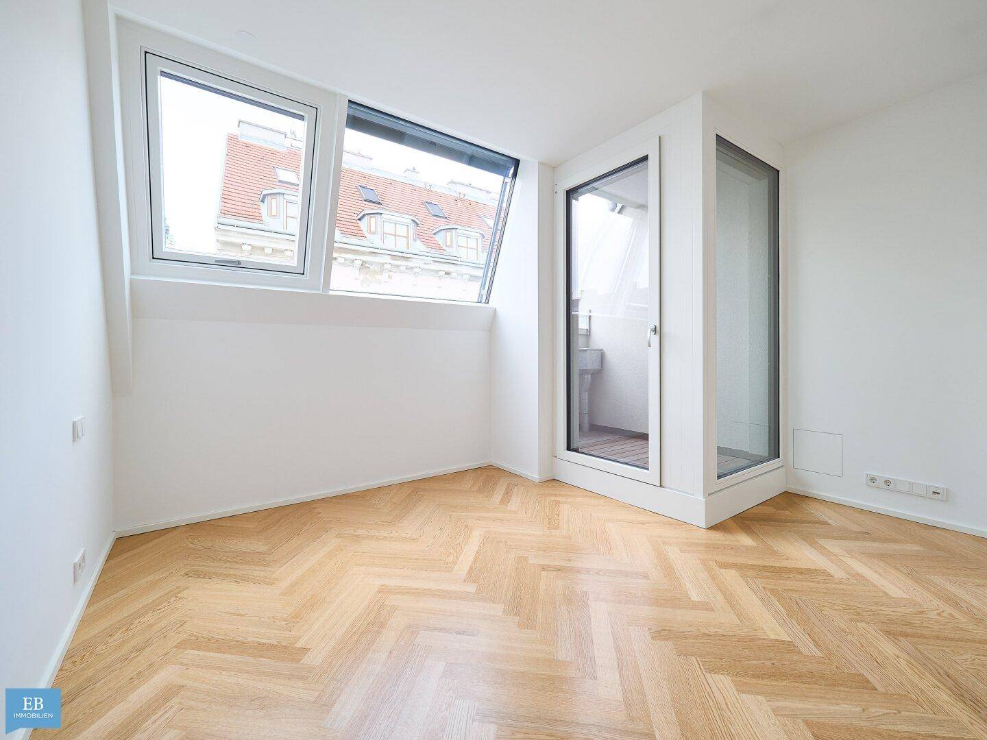 Lichtdurchflutetes Zimmer mit Fischgrätparkett, Dachfenster und Zugang zu einem schmalen Balkon.