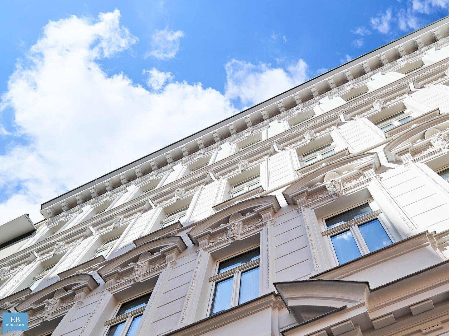 Historische Fassade eines Altbaus mit vielen Fenstern und dekorativen Elementen unter blauem Himmel.