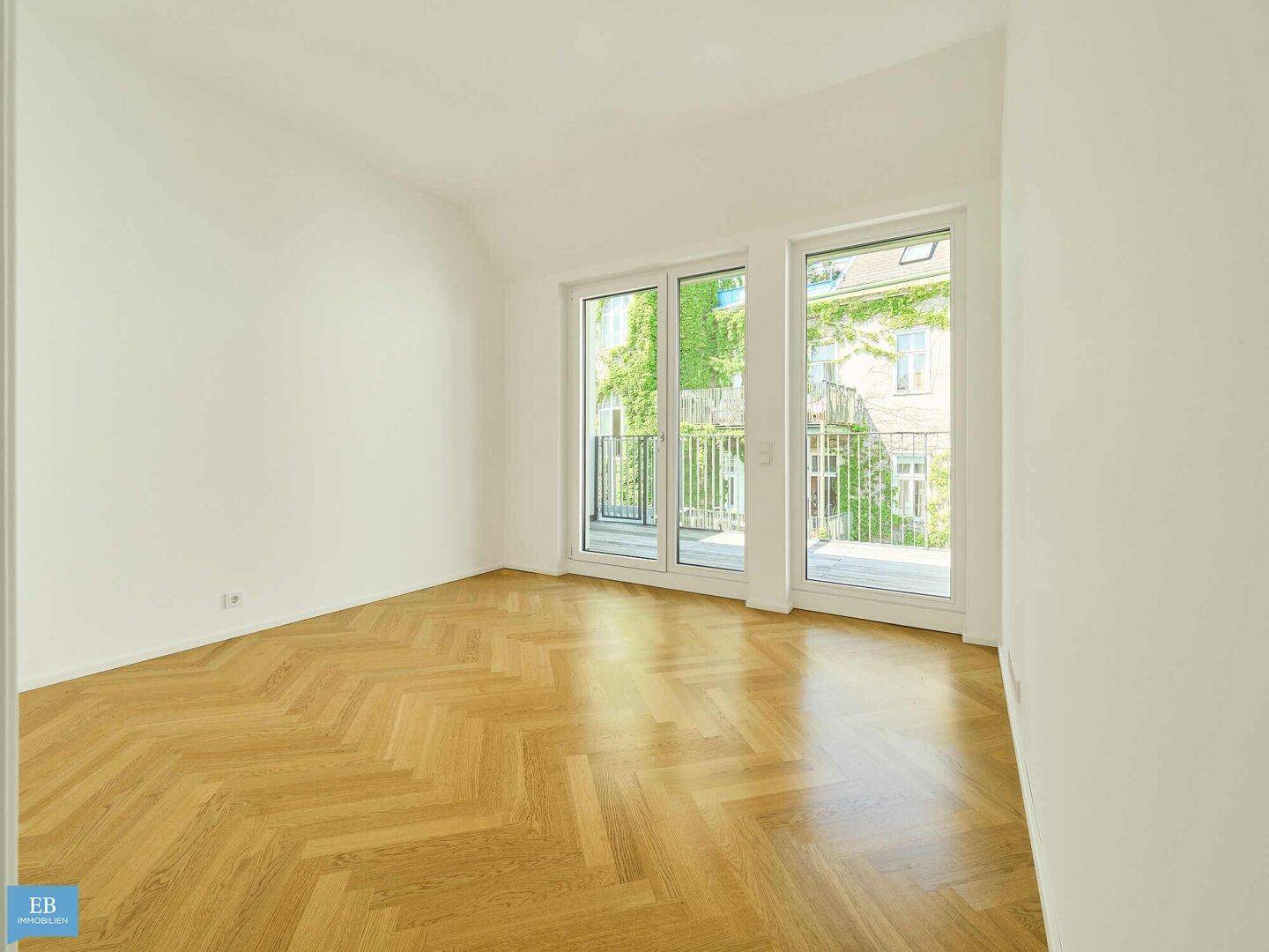 Geräumiger, heller Raum mit Fischgrätparkett und Zugang zu einem Balkon mit Grünblick.