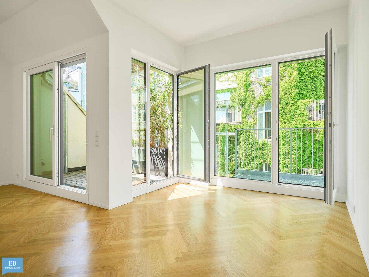 Lichtdurchfluteter Wohnraum mit Fischgrätparkett und großen Fenstern zum begrünten Balkon.