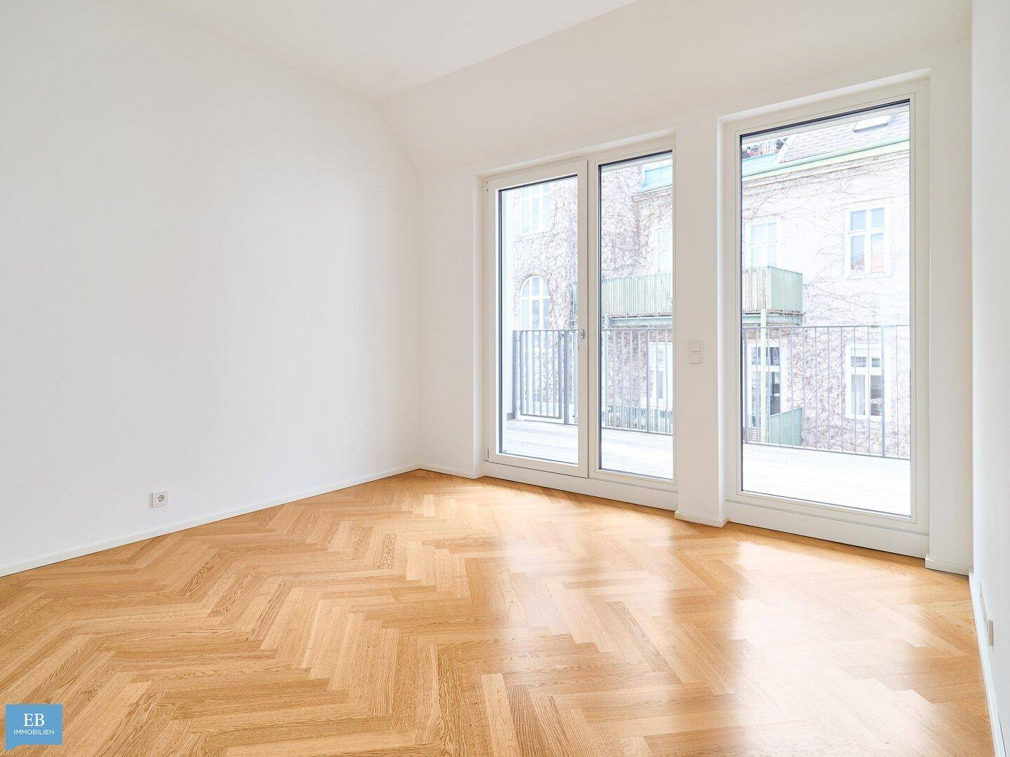Geräumiger, heller Wohnraum mit elegantem Fischgrätparkett und großen Fenstern zum Balkon.