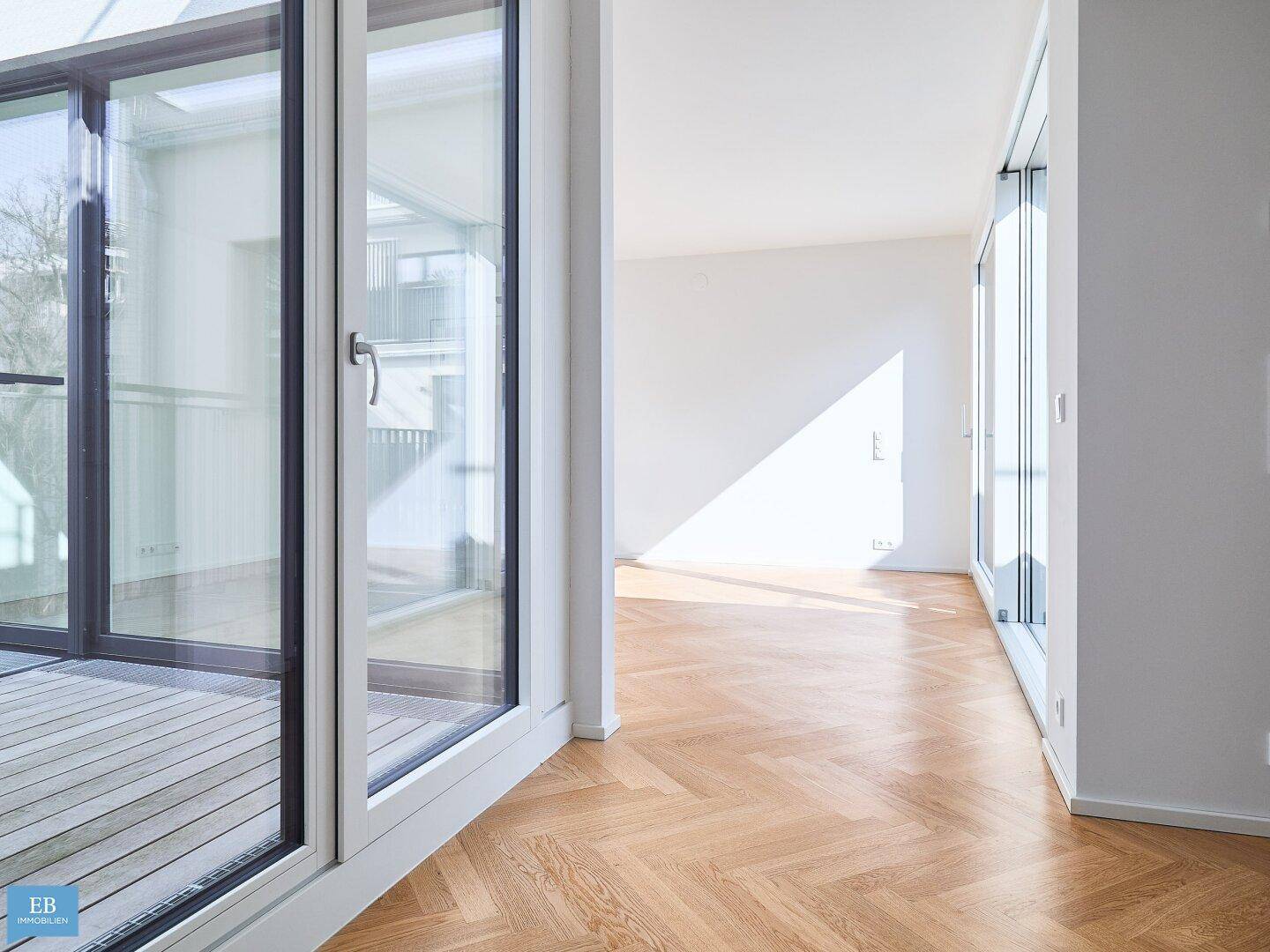 Heller Wohnraum mit Fischgrätparkett und breiten Fensterfronten, die Zugang zum Balkon ermöglichen.