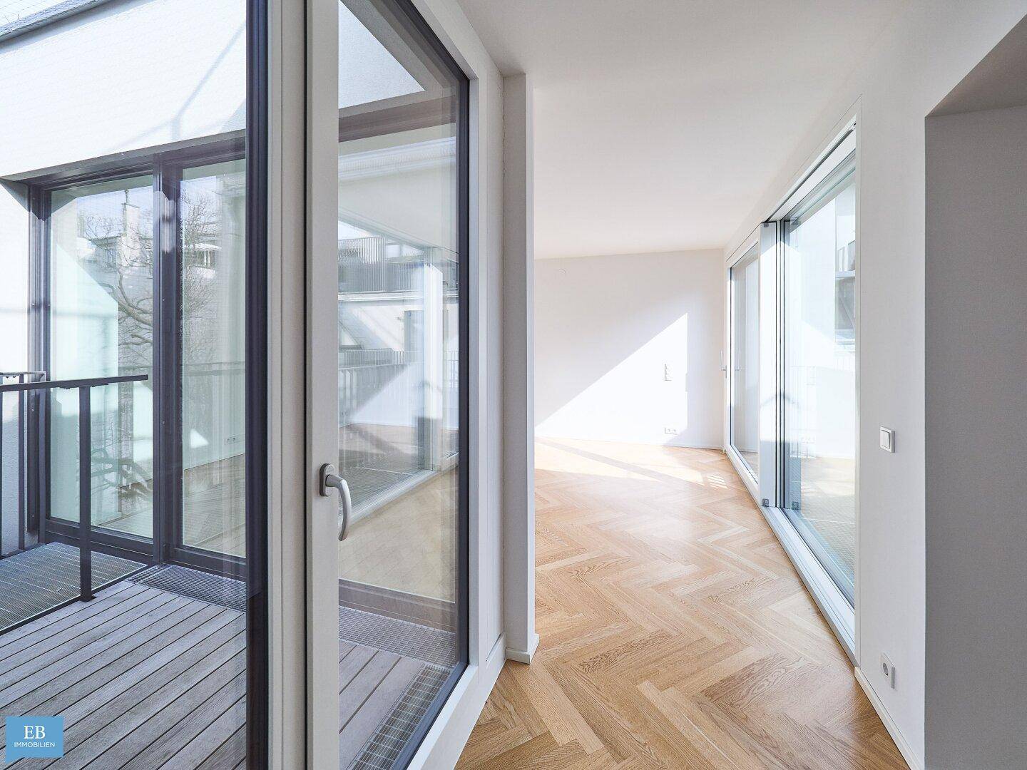 Heller Wohnraum mit Fischgrätparkett und breiten Fensterfronten, die Zugang zum Balkon ermöglichen.