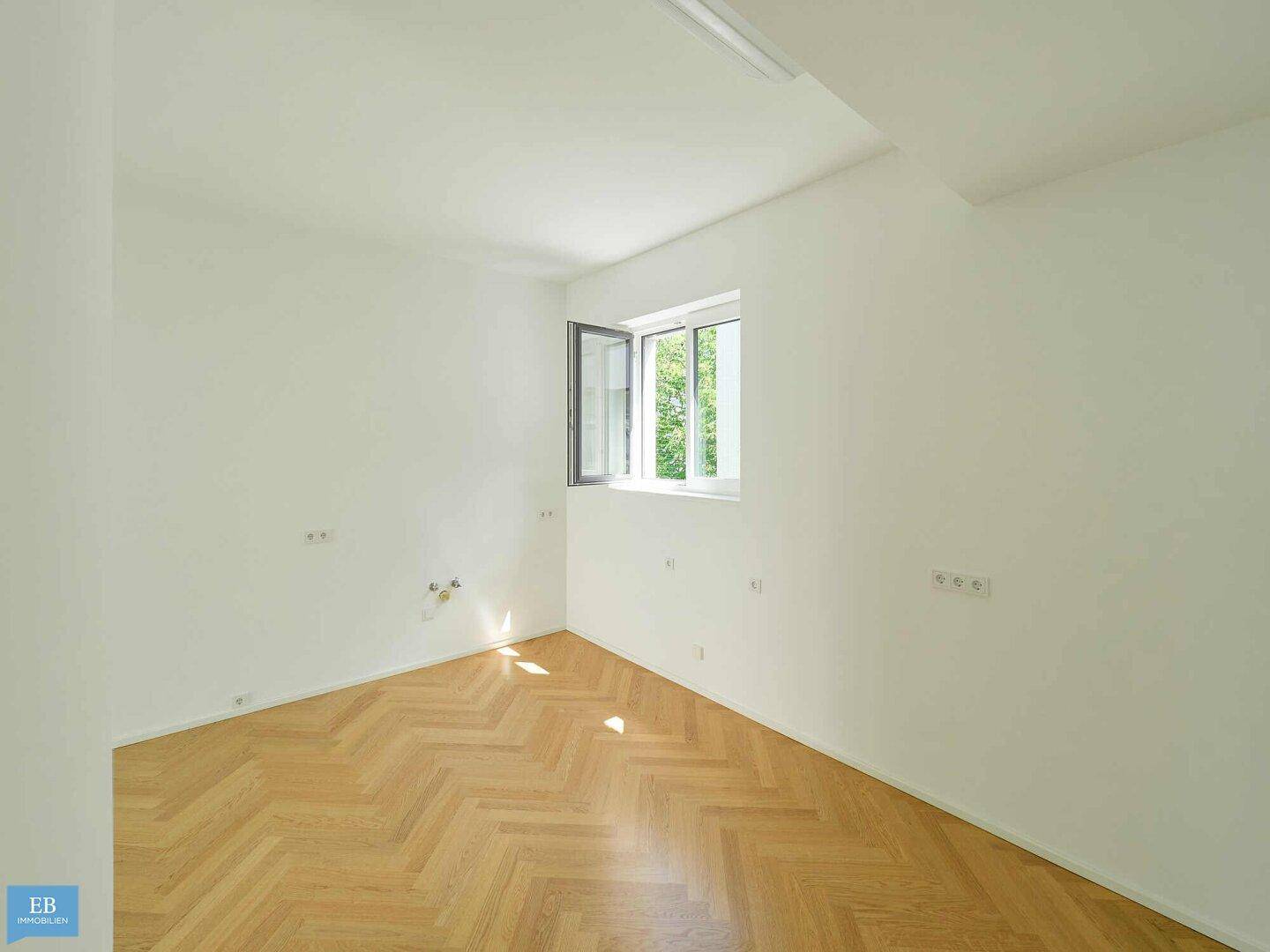 Helles Zimmer mit Fischgrätparkett und einem Fenster, das einen Blick ins Grüne bietet.