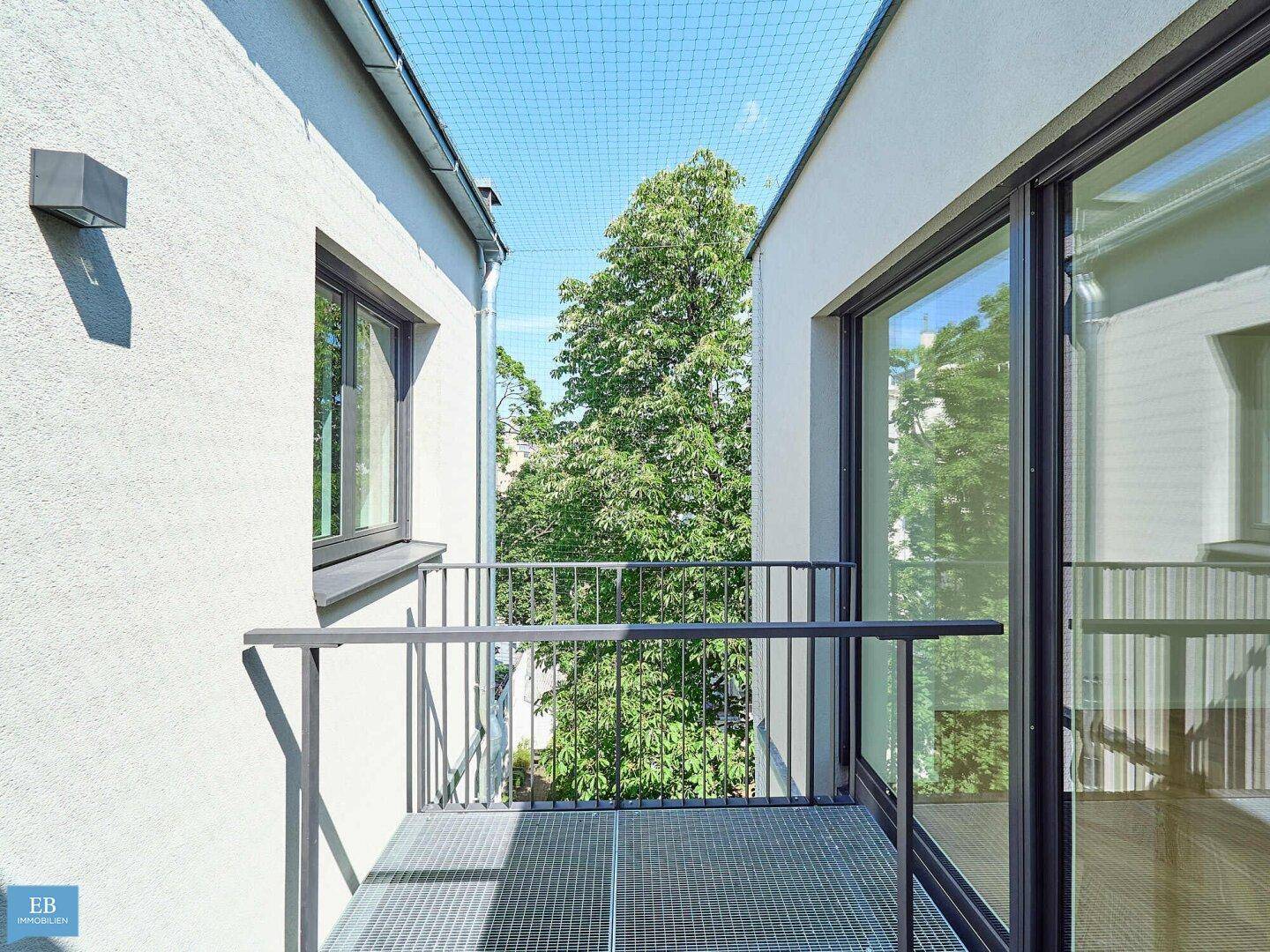 Kleiner Balkon mit Metallgeländer und Blick auf grüne Bäume, geschützt durch ein Netz.