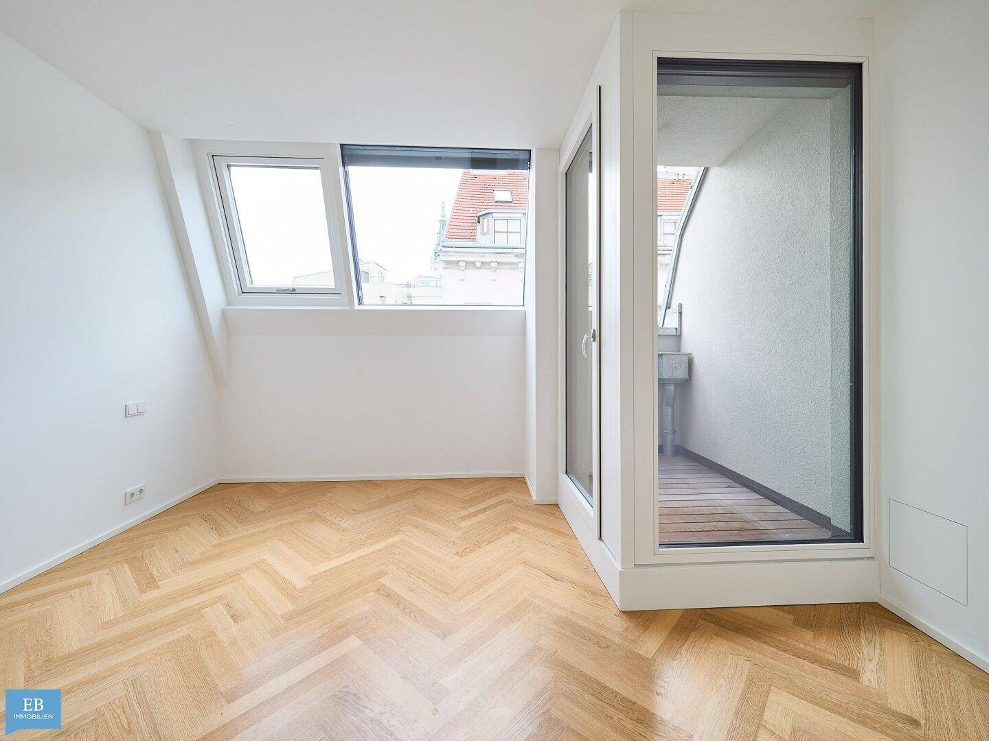 Helles Zimmer mit Fischgrätparkett, Dachfenster und Zugang zu einem kleinen, verglasten Balkon.