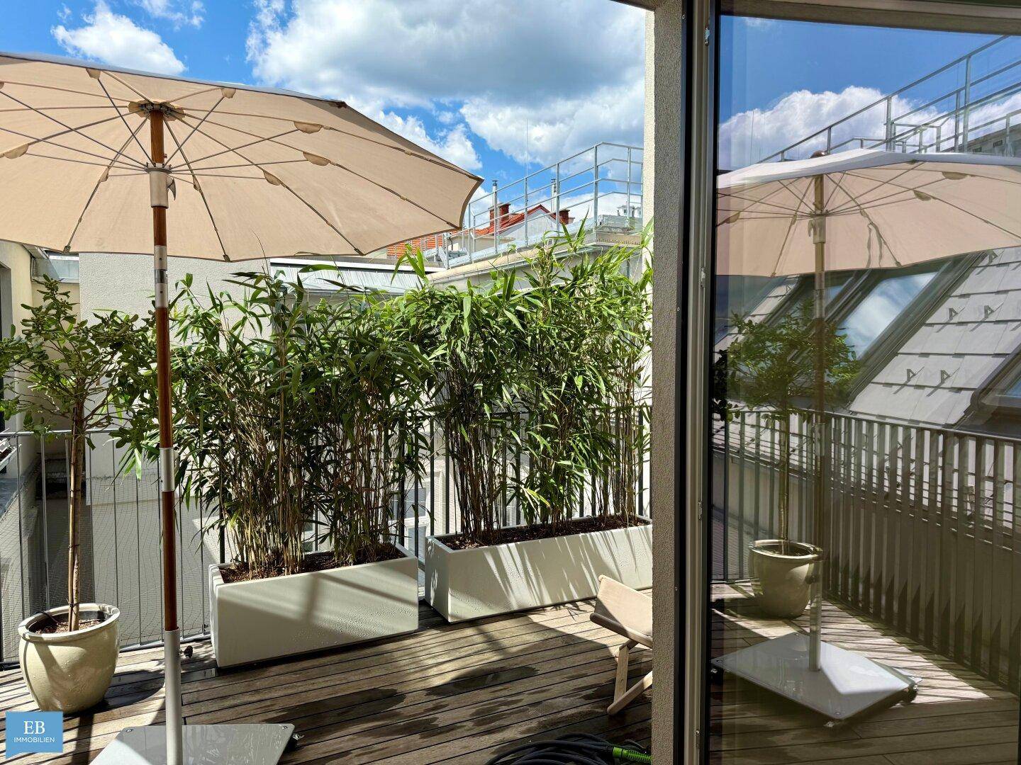 Geräumige Terrasse mit Holzboden, üppigen Bambuspflanzen und einem großen Sonnenschirm unter blauem Himmel.