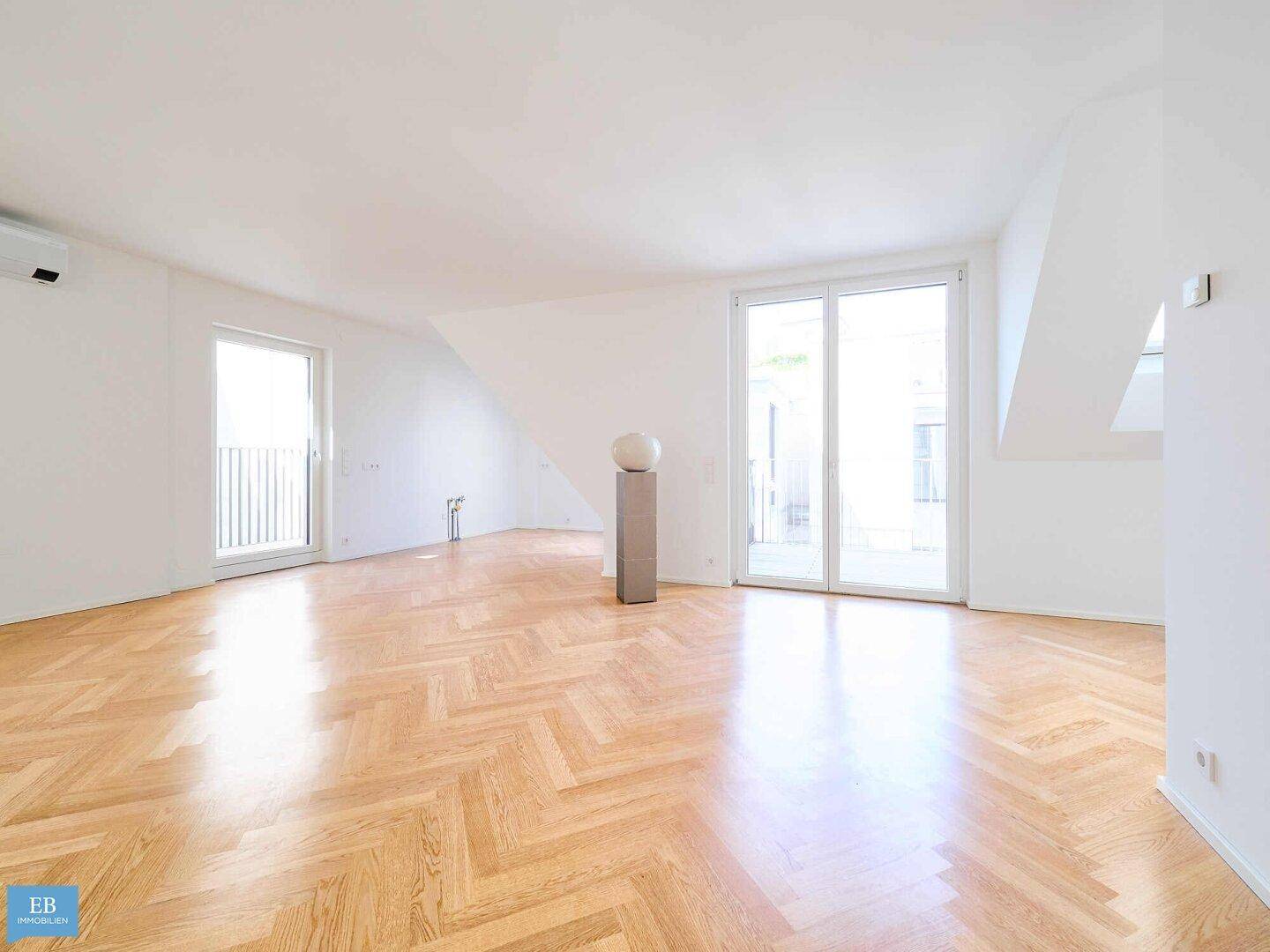 Großer, leerer Wohnraum mit elegantem Fischgrätparkett, weißen Wänden und Zugang zu mehreren Außenbereichen.