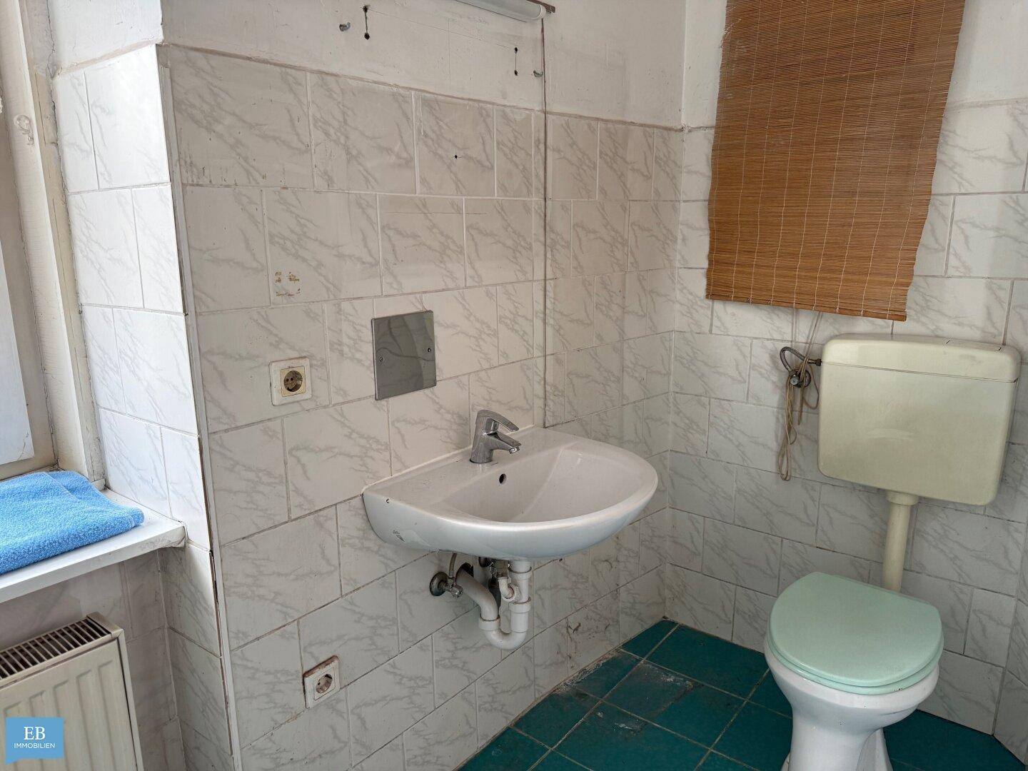 Badezimmer mit Toilette, Waschbecken und Fenster, gekennzeichnet durch grüne Boden- und weiße Wandfliesen.