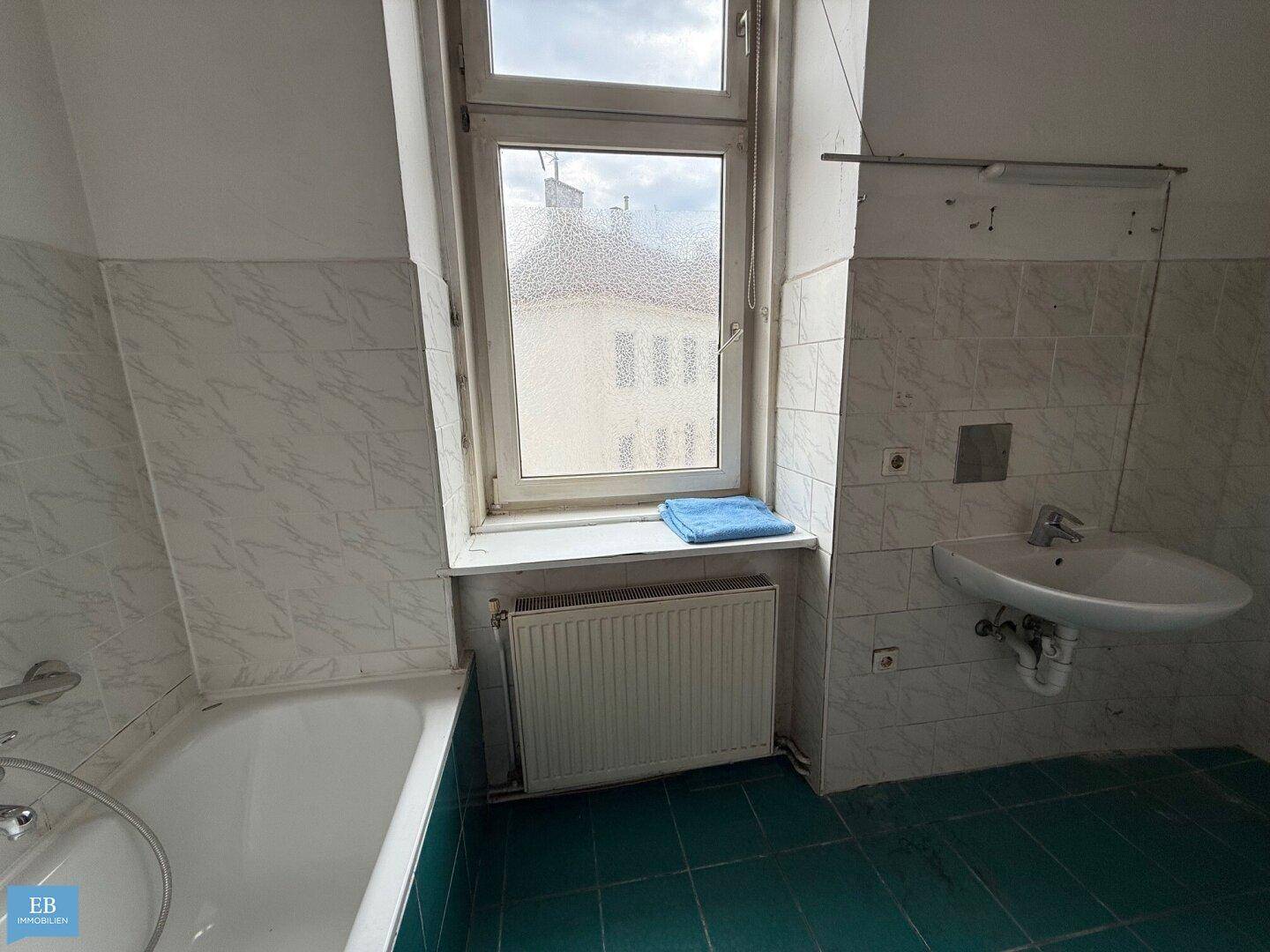 Badezimmer mit Badewanne, Waschbecken, Fenster und grünen Bodenfliesen, renovierungsbedürftig.