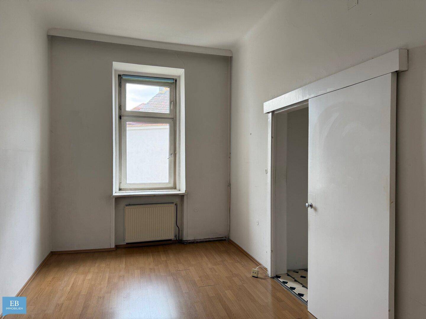 Helles Zimmer mit Holzboden, einem Fenster und einer Schiebetür, die zu einem gefliesten Bereich führt.