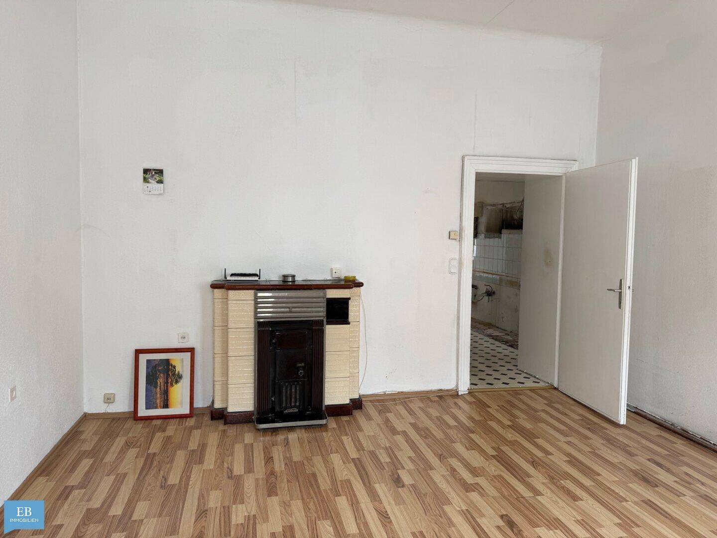 Heller Raum mit Holzboden und einem dekorativen Ofen, der zu einem gefliesten Bereich führt.