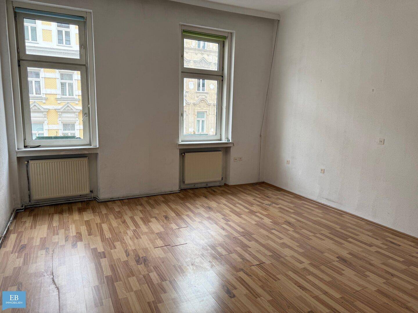Leeres Zimmer mit zwei großen Fenstern, Holzboden und Heizkörpern, die auf die Straße blicken.