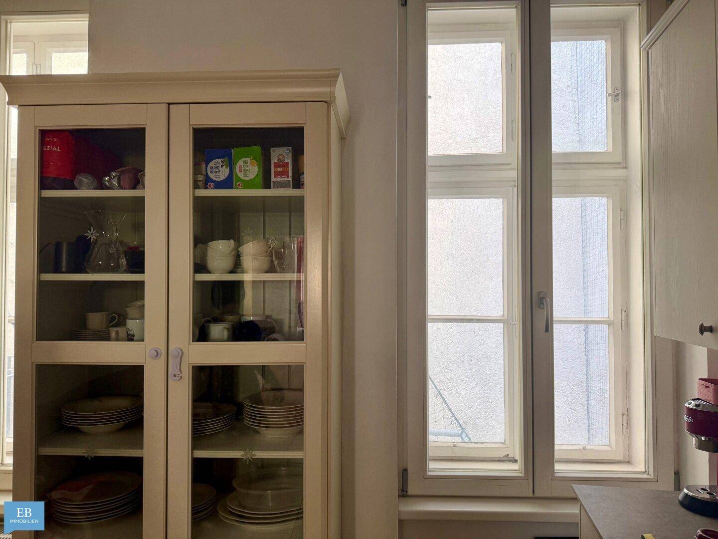 Heller Innenraum mit einem Geschirrschrank und hohen Fenstern, die viel Tageslicht hereinlassen.