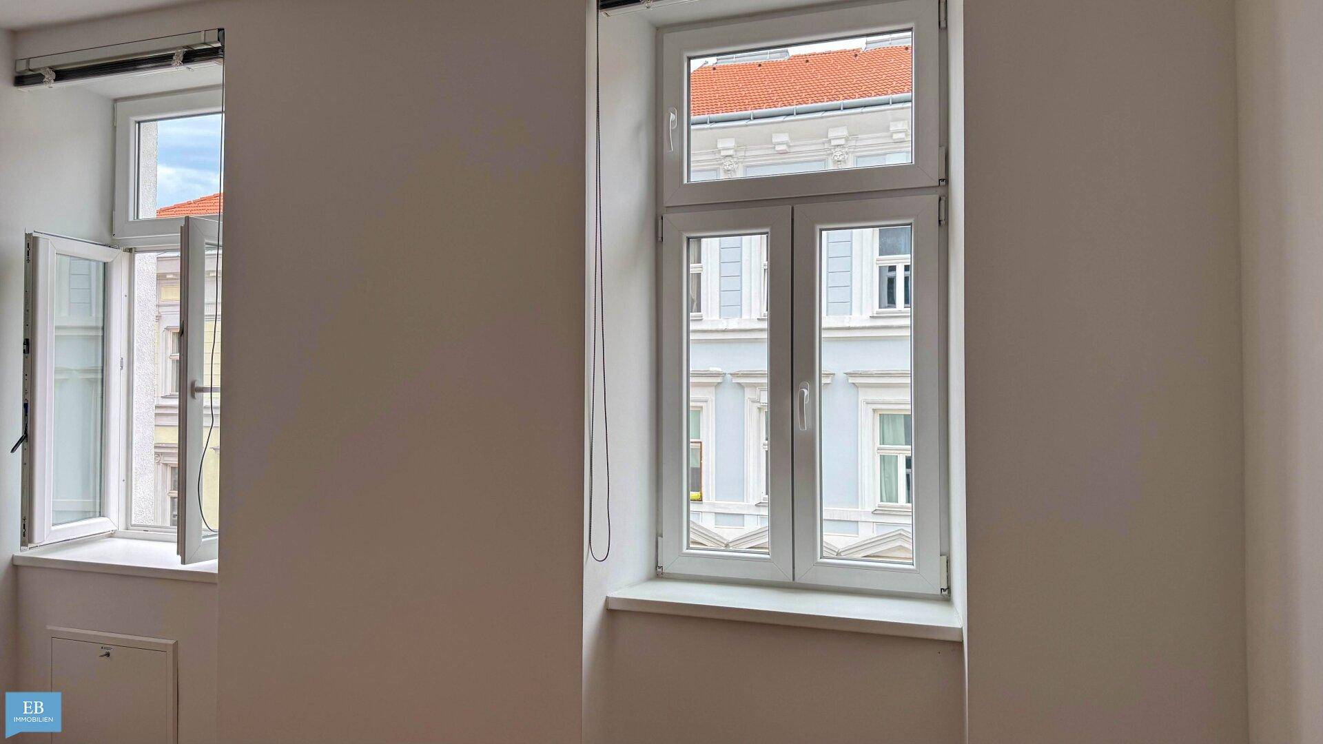 Heller Raum mit zwei Fenstern, eines davon geöffnet, bietet Blick auf die gegenüberliegende Fassade.