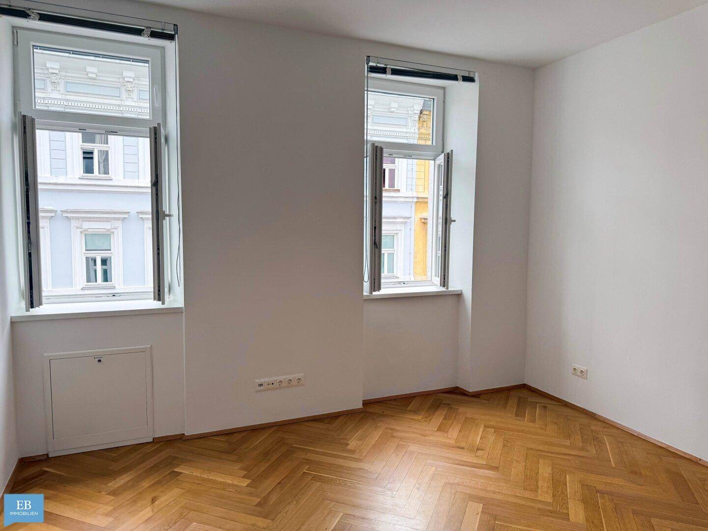 Heller Raum mit Fischgrätparkett und zwei Fenstern, die einen Blick auf die Stadt bieten.