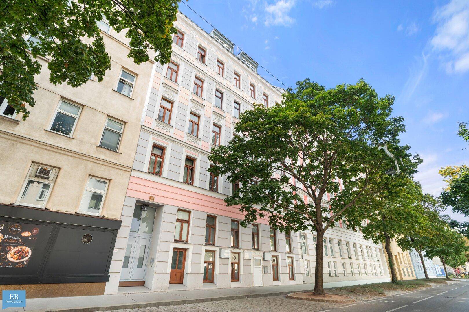 Historisches Gebäude mit klassischer Fassade und Bäumen an einer städtischen Straße.