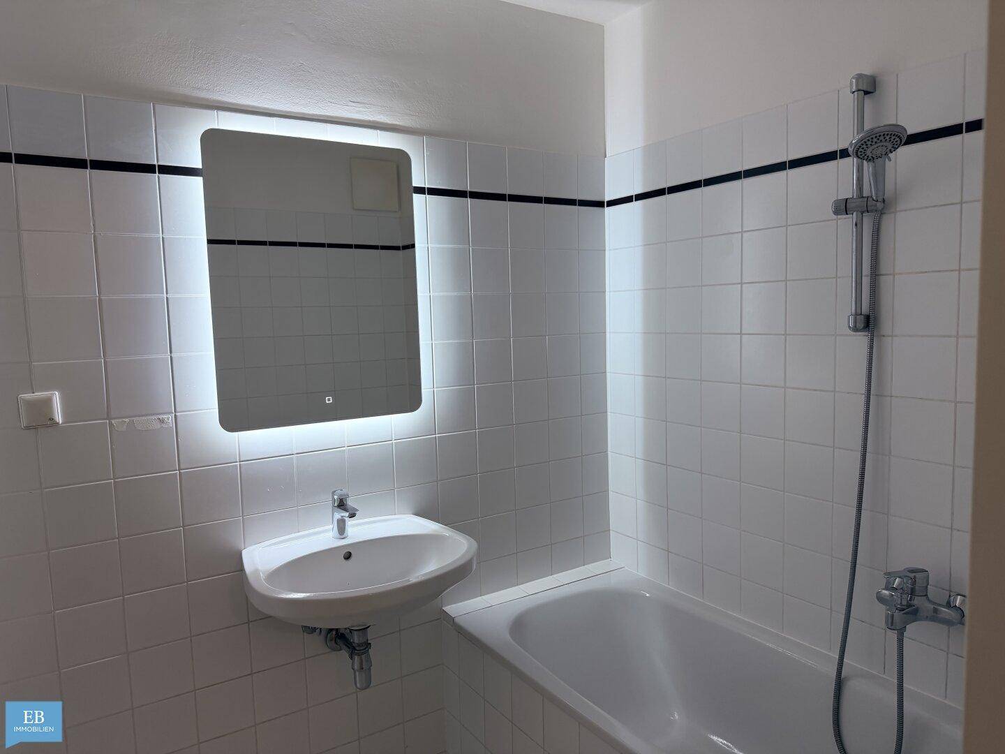 Helles Badezimmer mit weißen Fliesen, Badewanne und modernem beleuchtetem Spiegel.