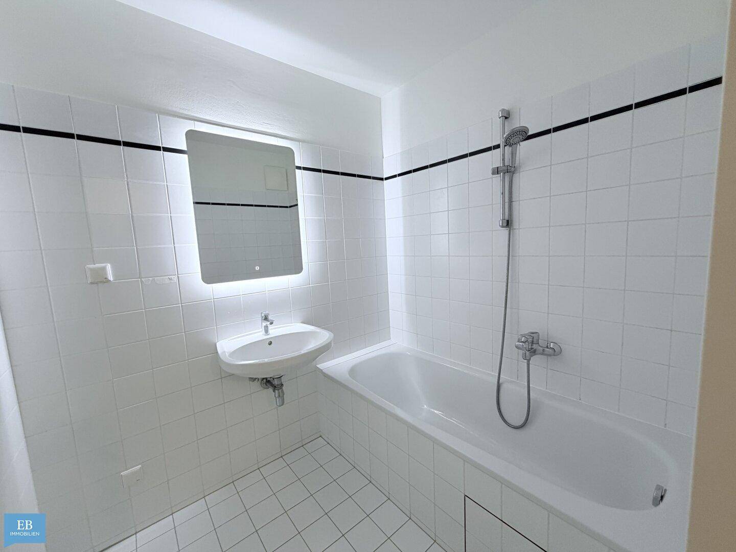 Modern gefliestes Badezimmer mit Badewanne, Waschbecken und beleuchtetem Spiegel.