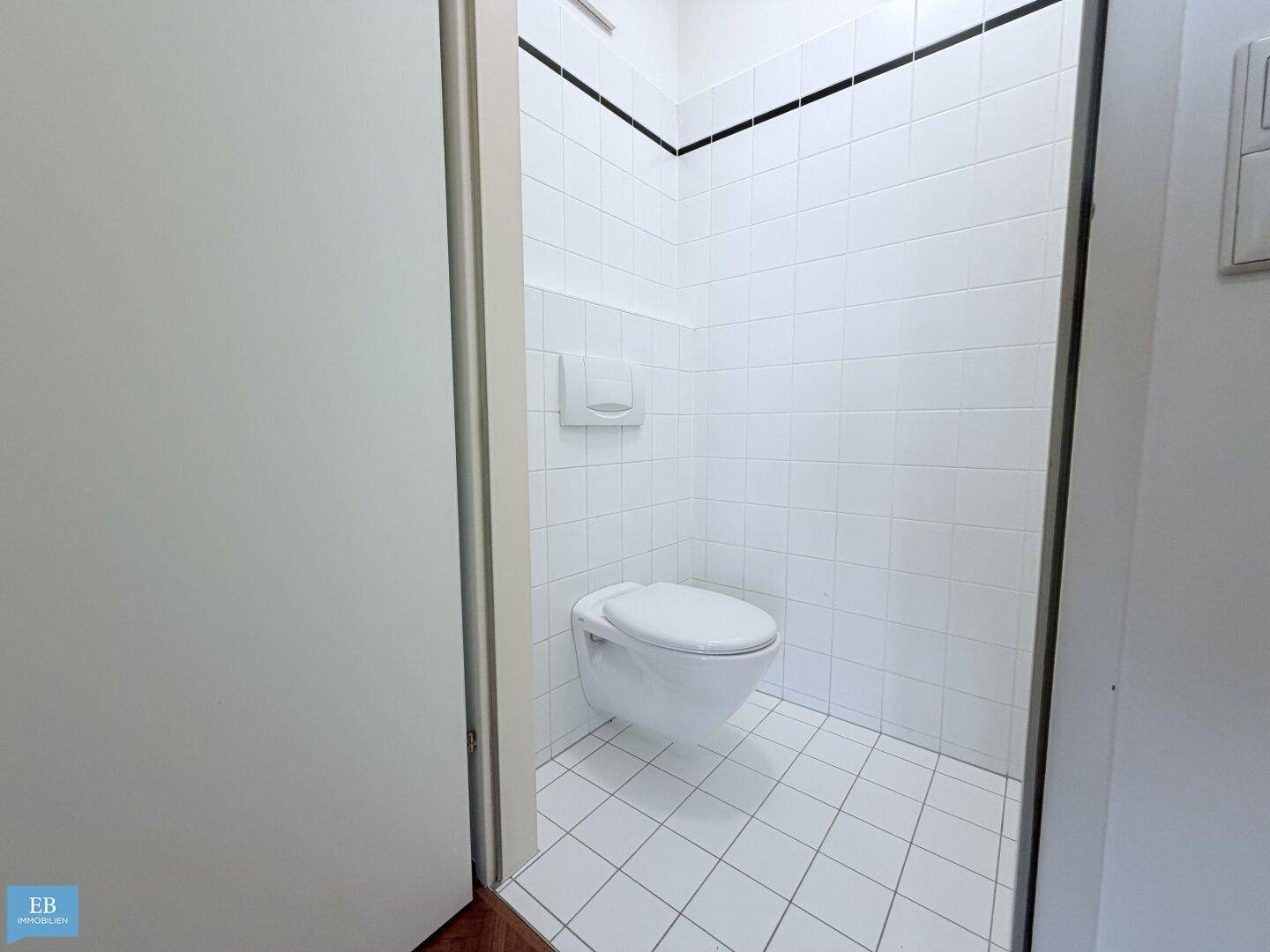 Separate Toilette mit wandhängendem WC und weißen Fliesen an Wänden und Boden.