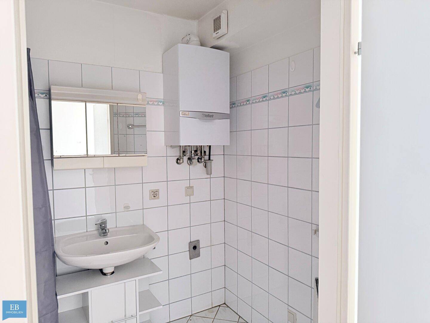 Badezimmer mit weißen Fliesen, Waschbecken, Spiegelschrank und Gastherme an der Wand.