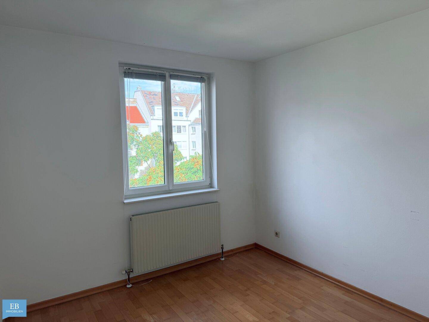 Leeres Schlafzimmer mit Parkettboden und einem Fenster, das viel Tageslicht hereinlässt.