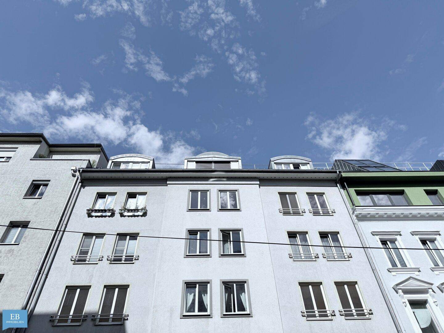 Fassade eines Mehrfamilienhauses mit mehreren Fenstern und Dachgauben unter blauem Himmel.
