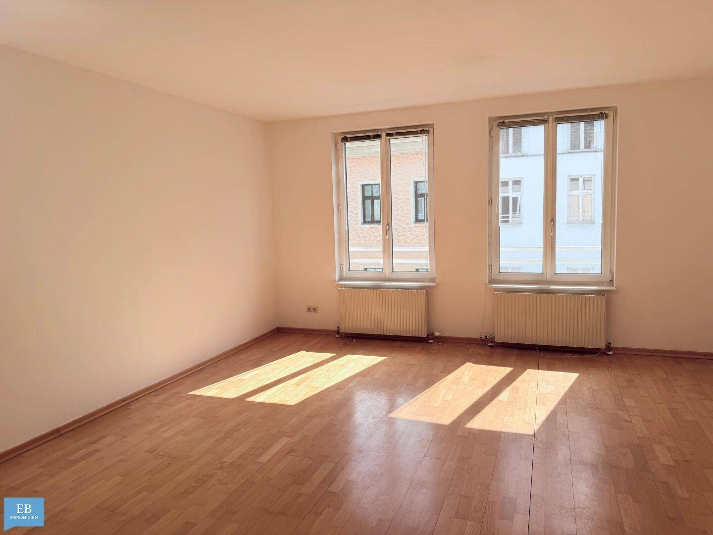 Helles, leeres Wohnzimmer mit Parkettboden und zwei großen Fenstern, durch die Sonnenlicht fällt.