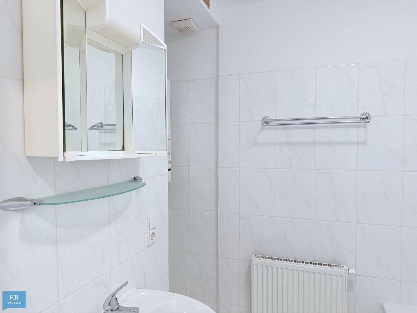 Helles Badezimmer mit weißen Fliesen, Waschbecken, Spiegelschrank und Heizkörper.
