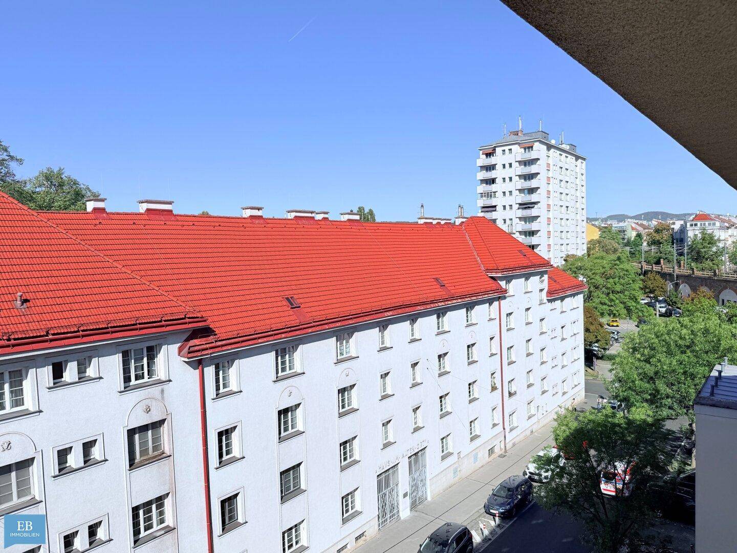 Blick auf ein Wohngebäude mit rotem Ziegeldach und einer belebten Straße mit parkenden Autos.
