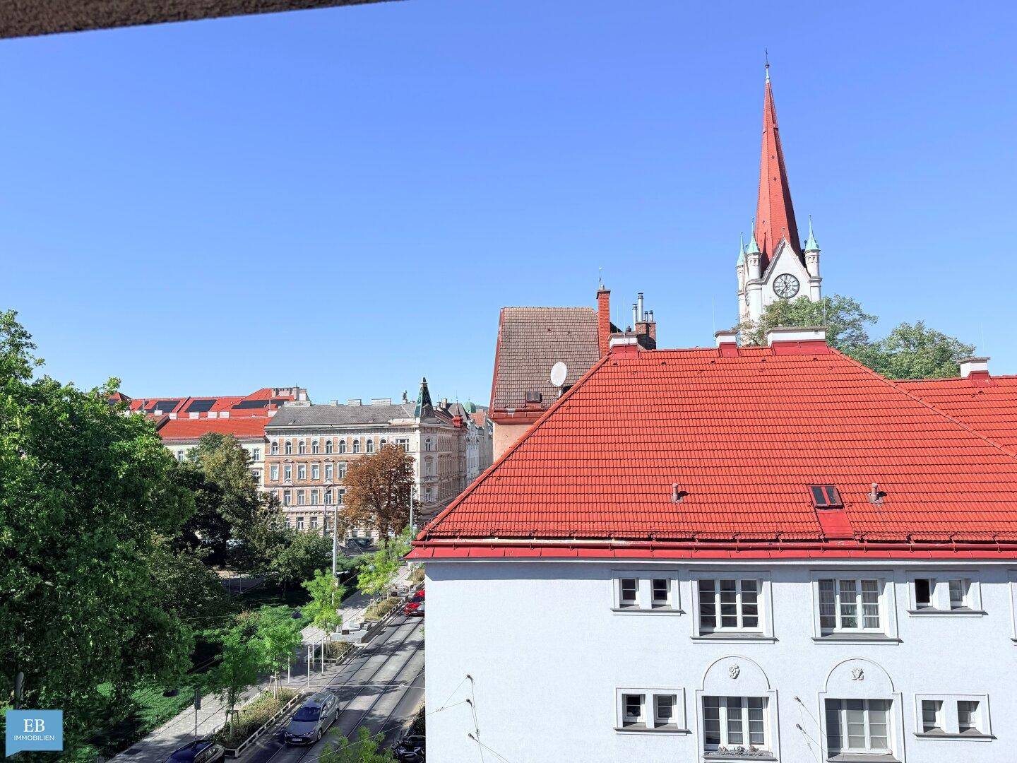Stadtansicht mit einem Gebäude mit rotem Ziegeldach und einem Kirchturm im Hintergrund unter blauem Himmel.