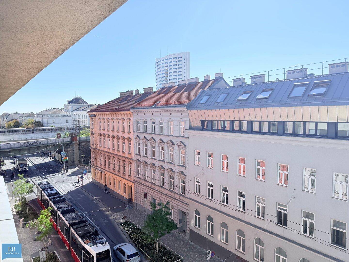Panoramablick auf eine städtische Straße mit historischen Gebäuden, einer Straßenbahnlinie und einem modernen Hochhaus im Hintergrund.