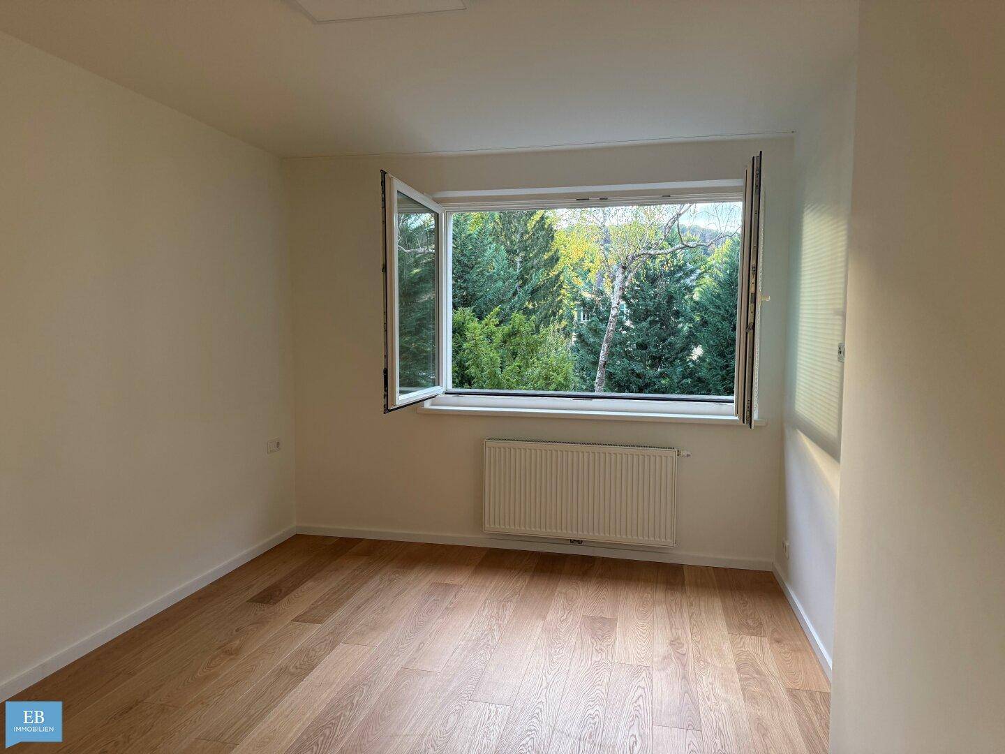 Helles Schlafzimmer mit Holzboden und einem geöffneten Fenster mit Blick ins Grüne.