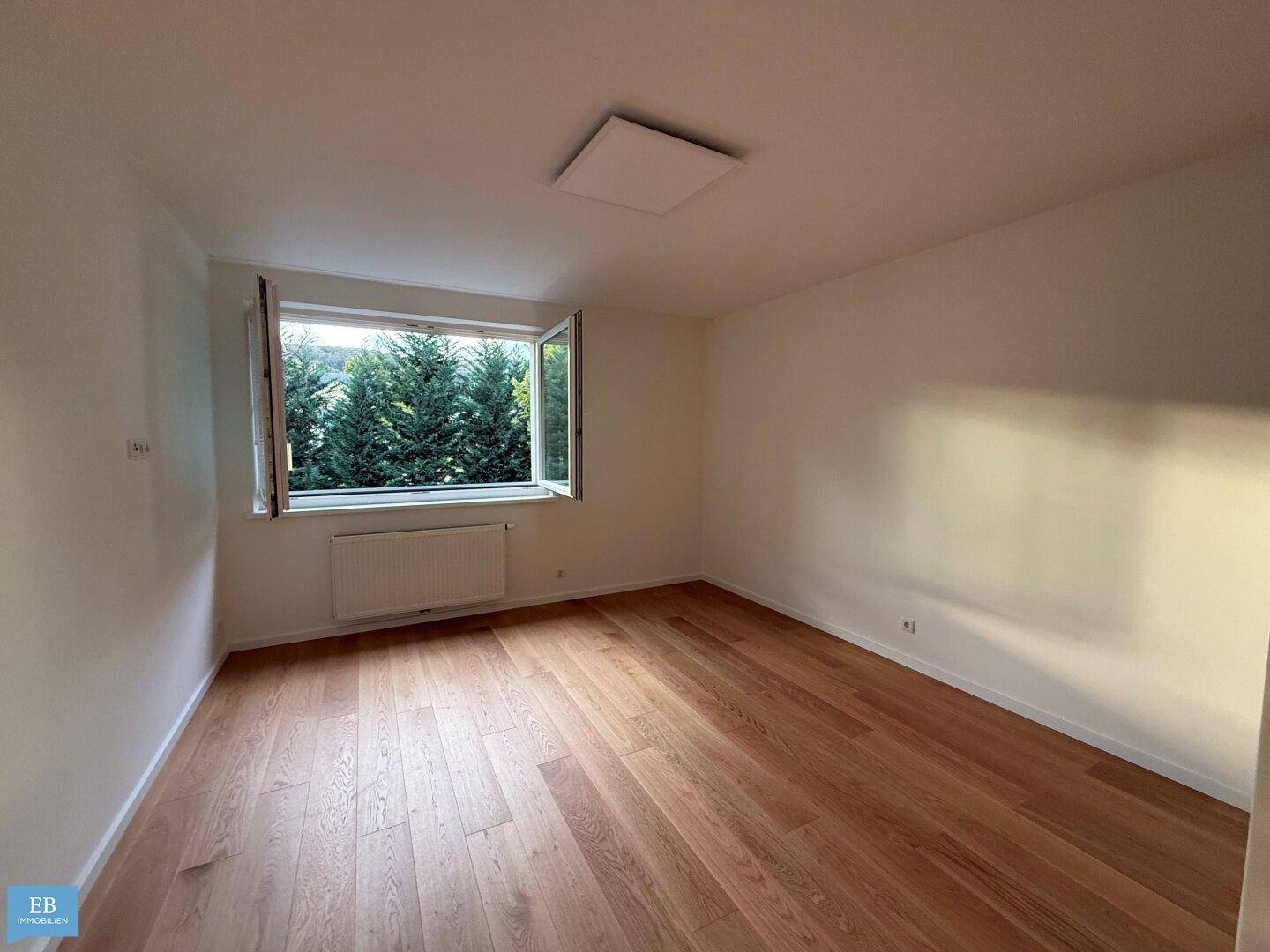Geräumiges Zimmer mit Holzboden, weißen Wänden und einem Fenster mit Blick auf Bäume.