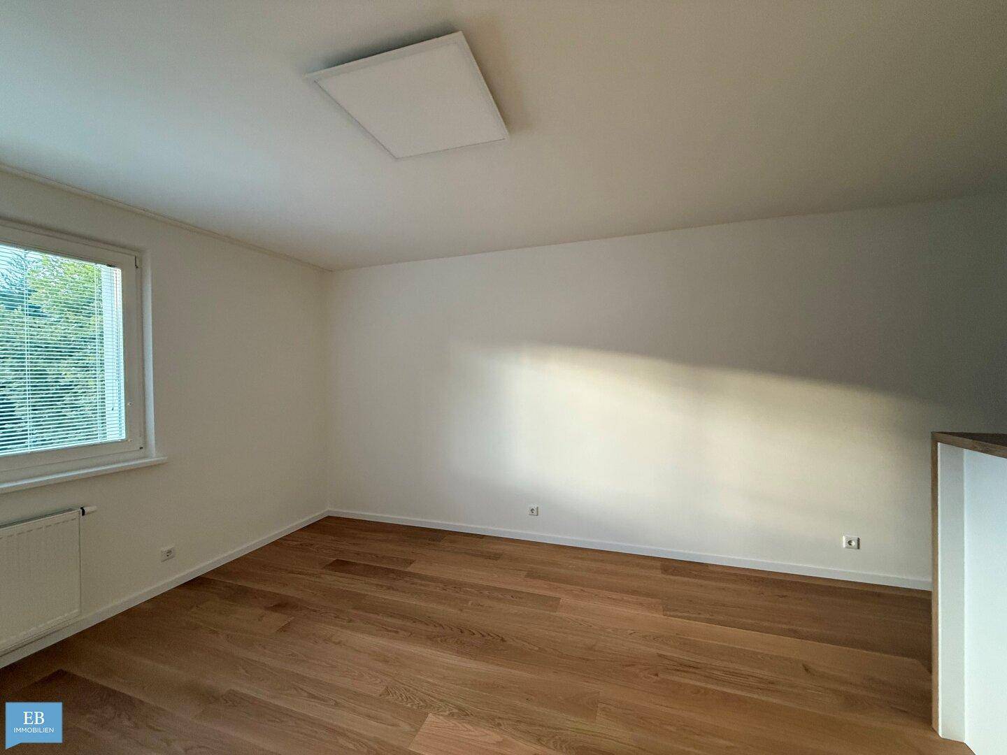 Leeres Zimmer mit Holzboden, weiß gestrichenen Wänden und einem Fenster mit Jalousien.