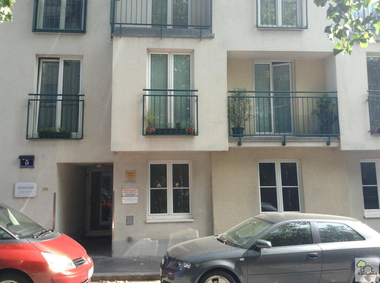 Fassade eines Mehrfamilienhauses mit mehreren Balkonen und geparkten Autos davor.