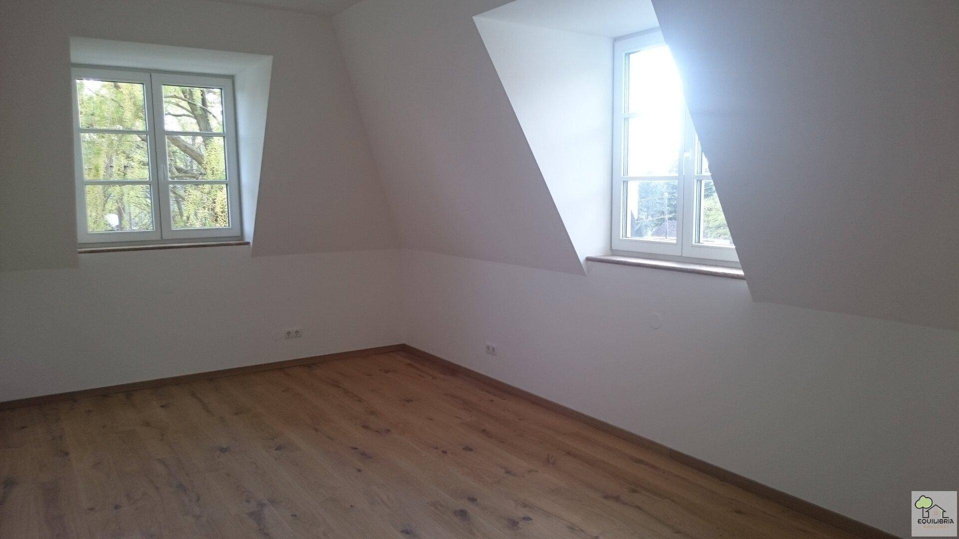 Dachgeschosszimmer mit schrägen Wänden, Holzfußboden und zwei Fenstern für viel Licht.