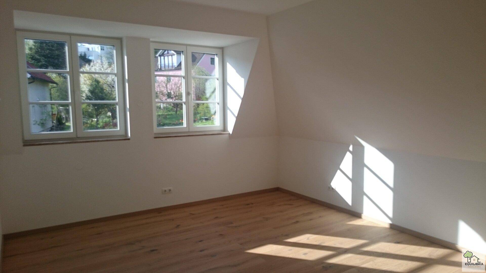 Sonnenverwöhntes Zimmer mit Holzboden und Blick ins Grüne durch zwei Fenster.