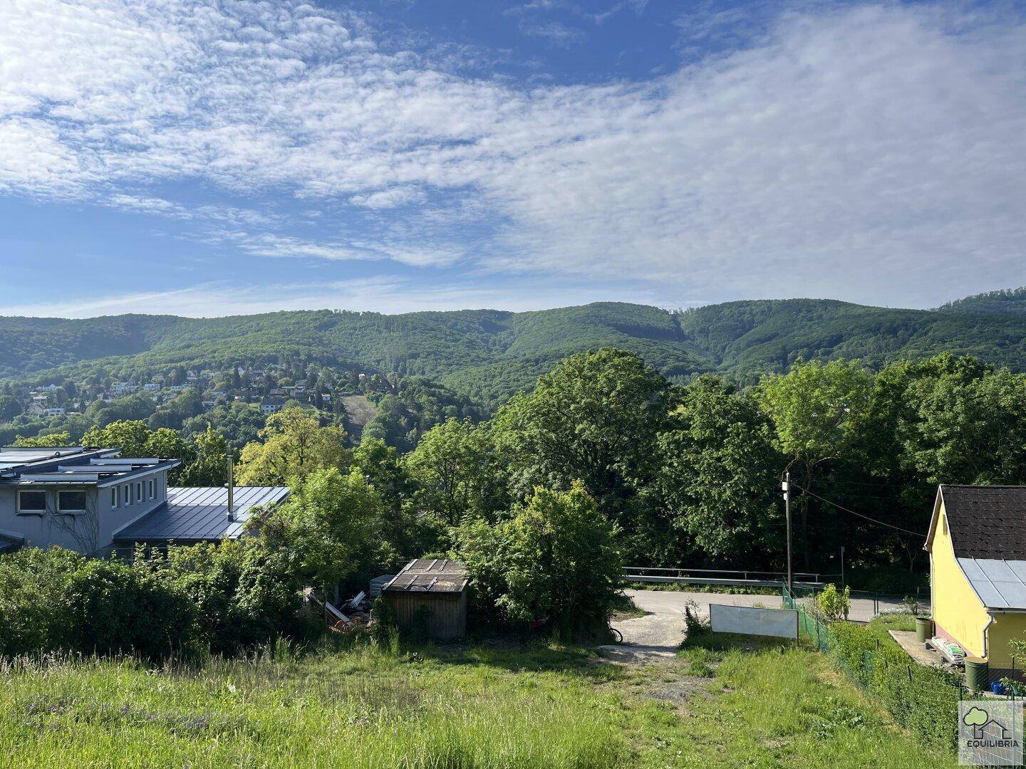 Weitläufiger Blick über eine grüne Landschaft mit Bäumen, Häusern und bewaldeten Hügeln.