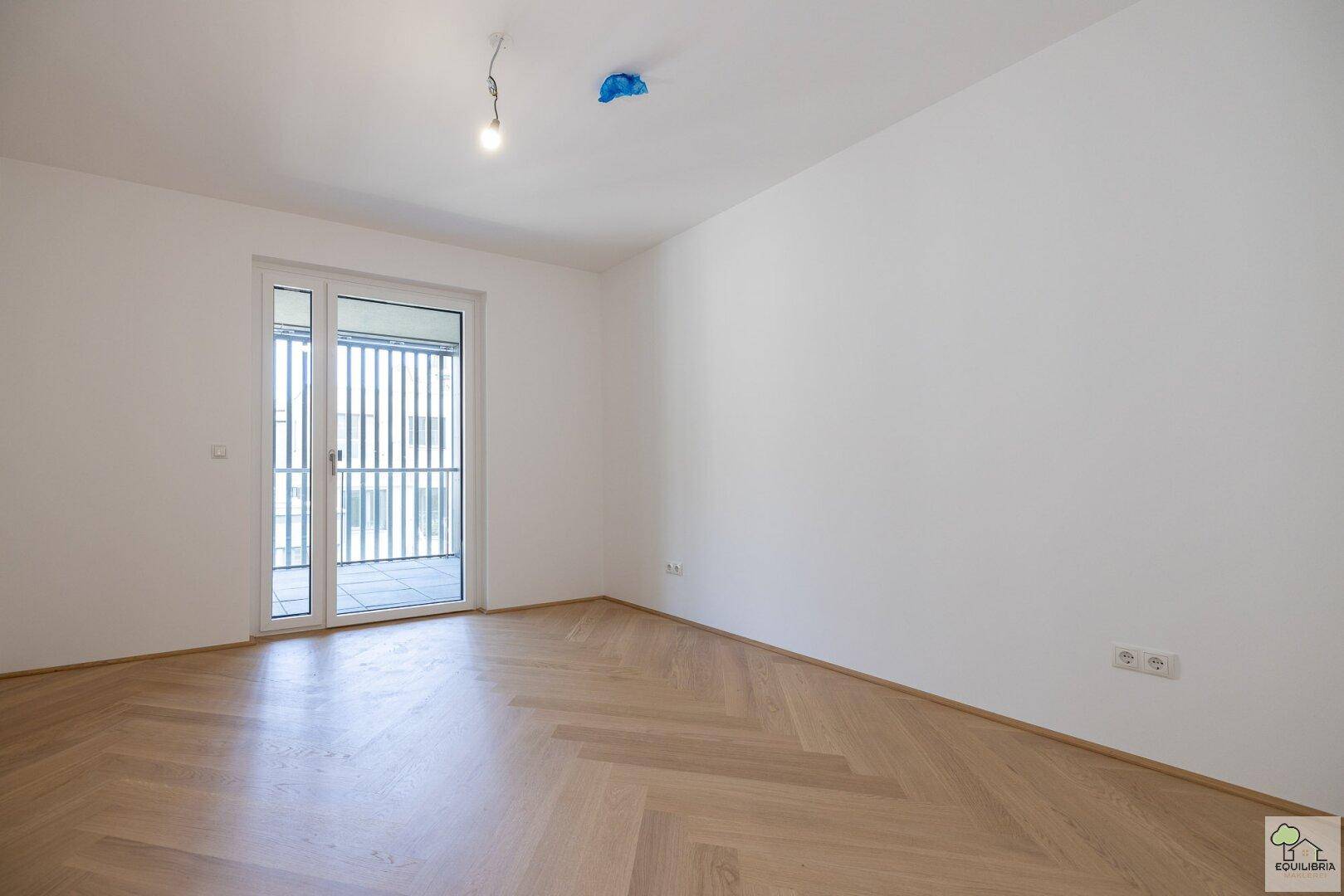 Leeres Zimmer mit Fischgrätparkett und großem Fenster, das Zugang zu einem französischen Balkon bietet.