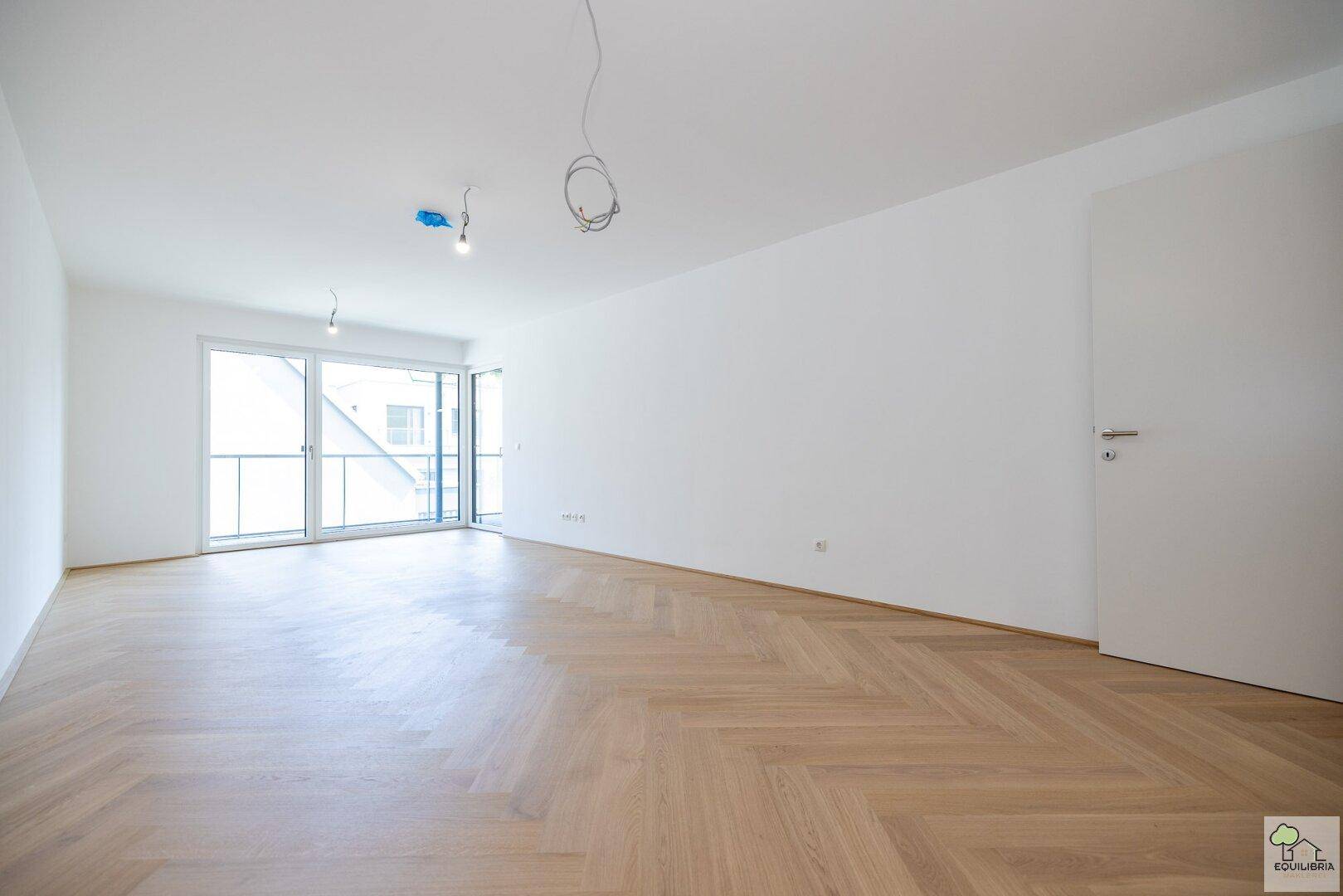 Geräumiges, helles Wohnzimmer mit Fischgrätparkett und breiter Fensterfront zum Außenbereich.