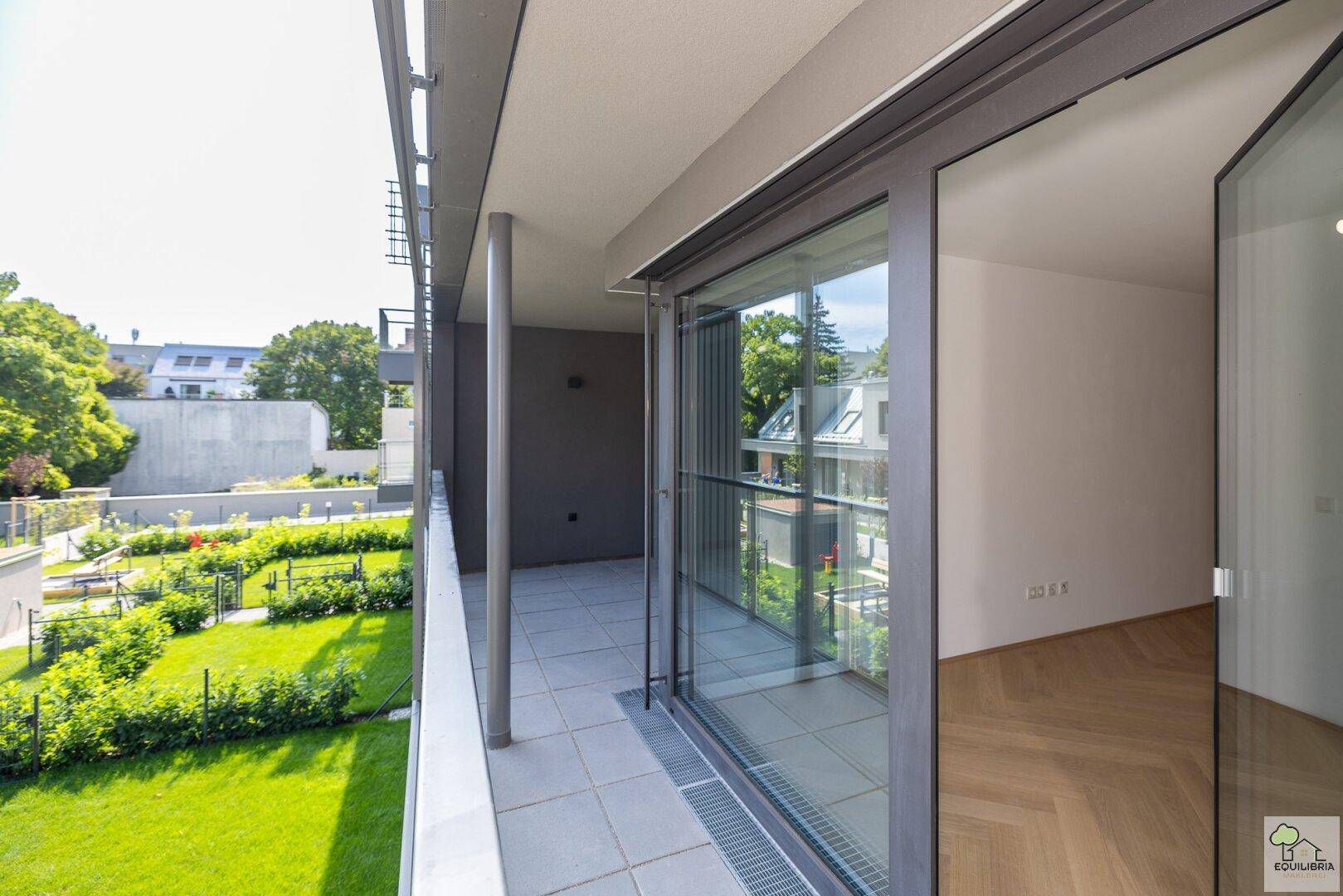 Geräumiger Balkon mit Fliesenboden und Glasgeländer, bietet Blick auf den grünen Gartenbereich.