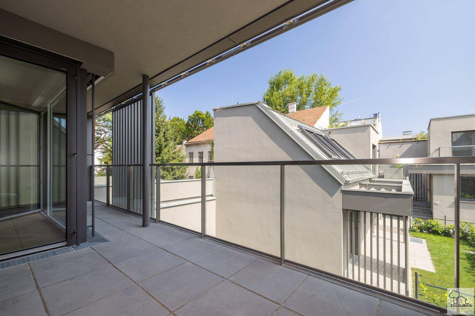 Moderner Balkon mit Fliesenboden und Glasgeländer, umgeben von grünen Anlagen und Gebäuden.