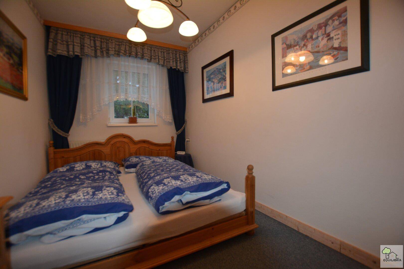 Schlafzimmer mit einem traditionellen Holzbett, Teppichboden und einem kleinen Fenster mit Vorhängen.