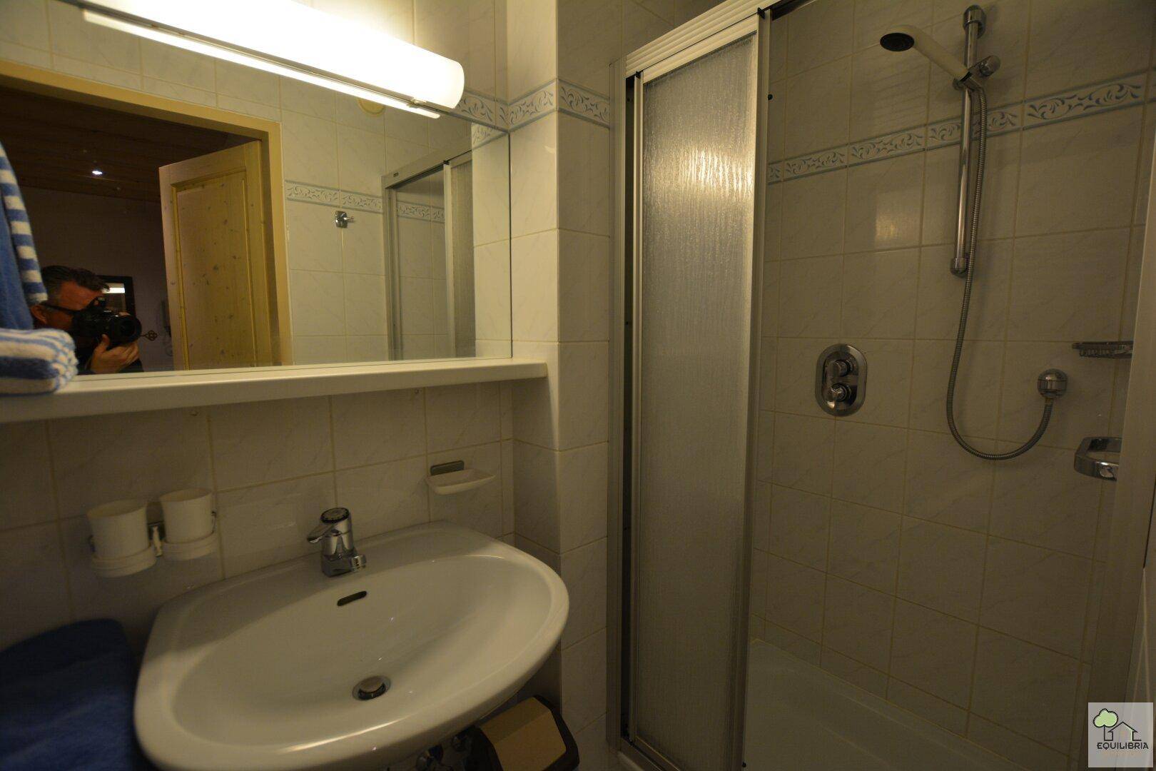 Helles Badezimmer mit einer Duschkabine, Waschbecken und weißen Fliesen an den Wänden.