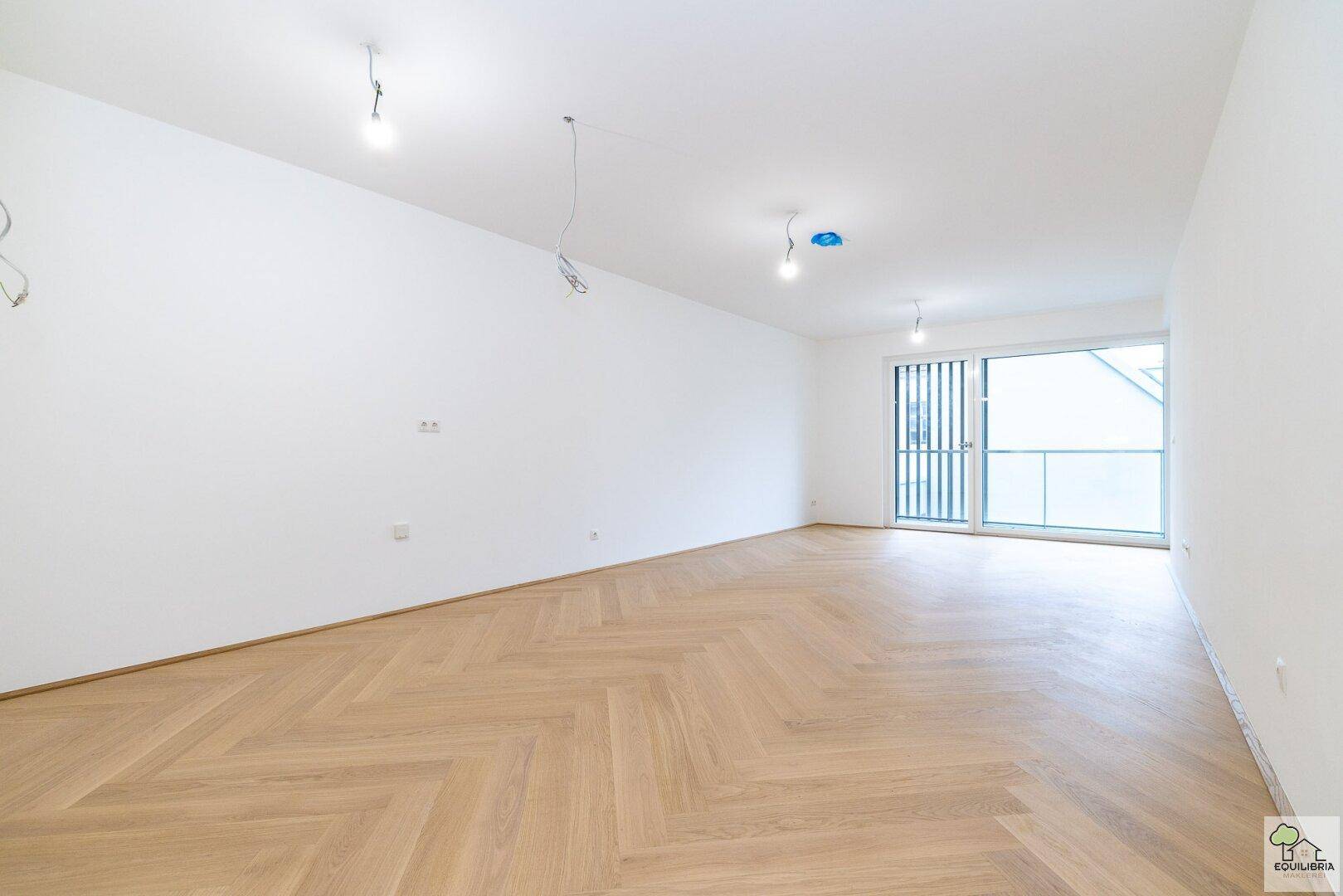 Geräumiger Wohnbereich mit hellem Fischgrätparkett und großem Fenster zum Balkon.
