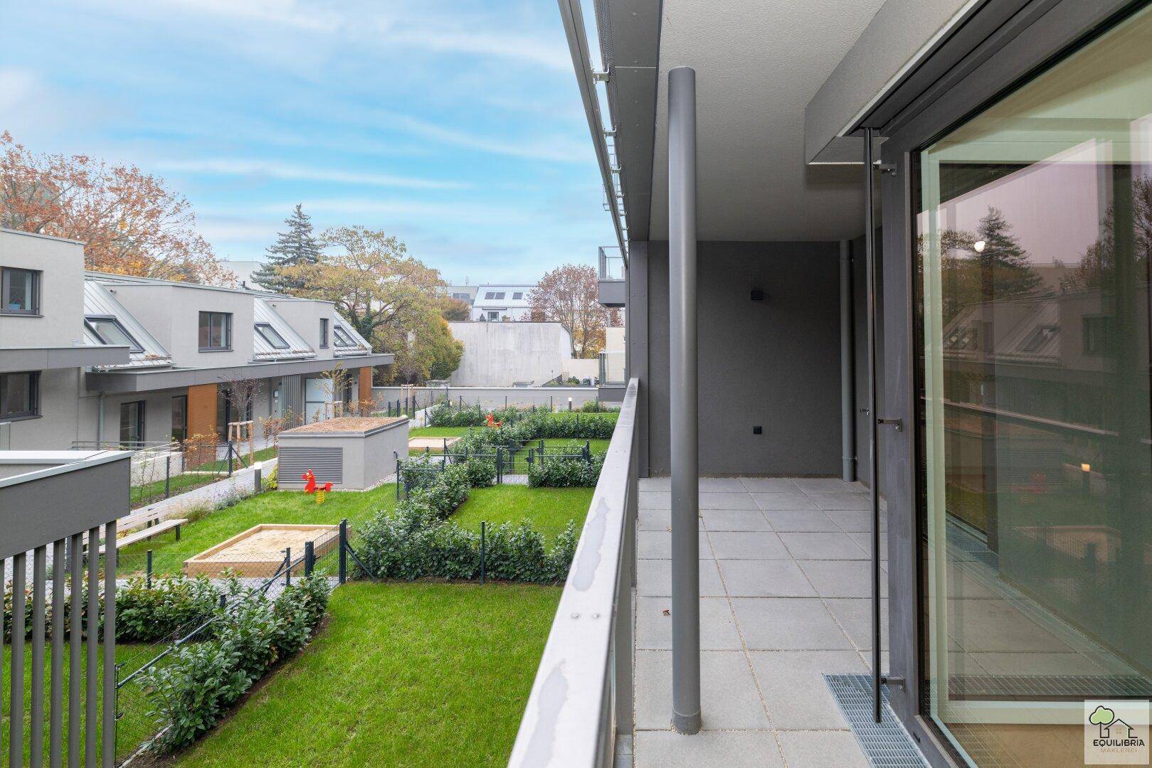 Sonniger Balkon mit weitem Blick auf den grünen Gemeinschaftsgarten und Spielplatz.