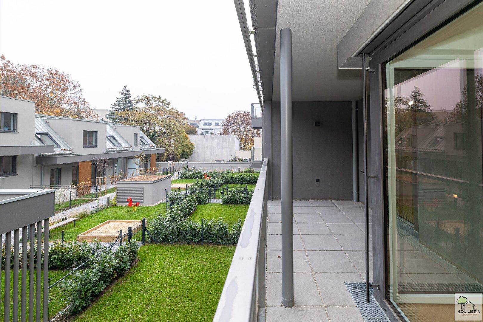 Moderner Balkon mit Fliesenboden und Blick auf den gepflegten Gemeinschaftsgarten mit Spielplatz und Hecken.