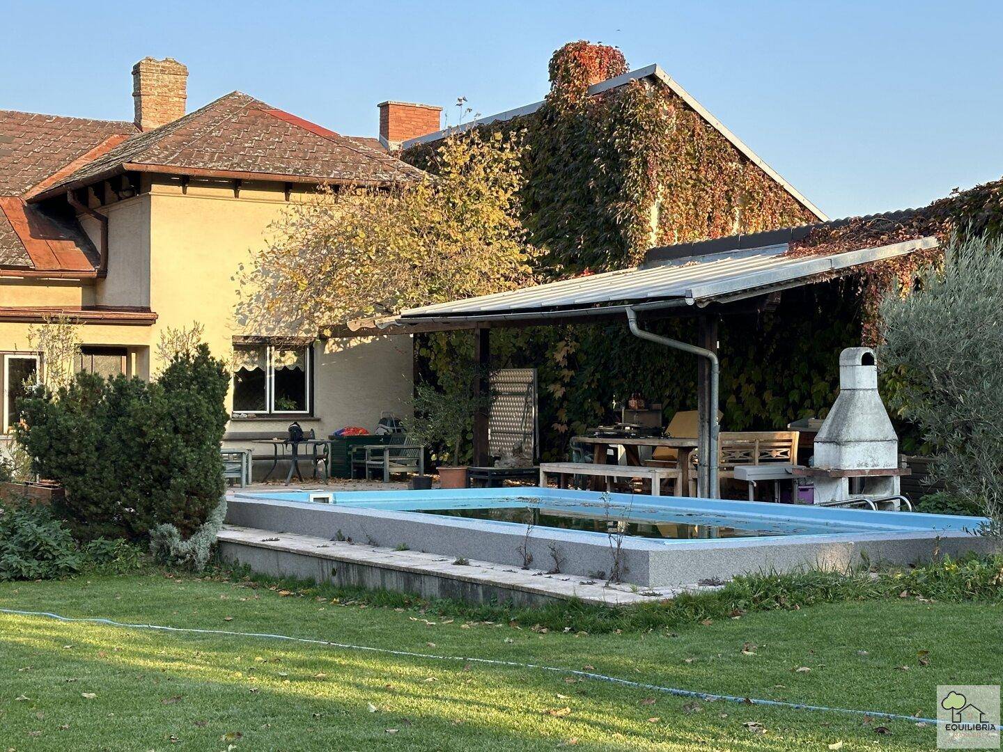 Außenbereich mit Pool, überdachtem Sitzplatz und gemauertem Grill neben dem Haus.