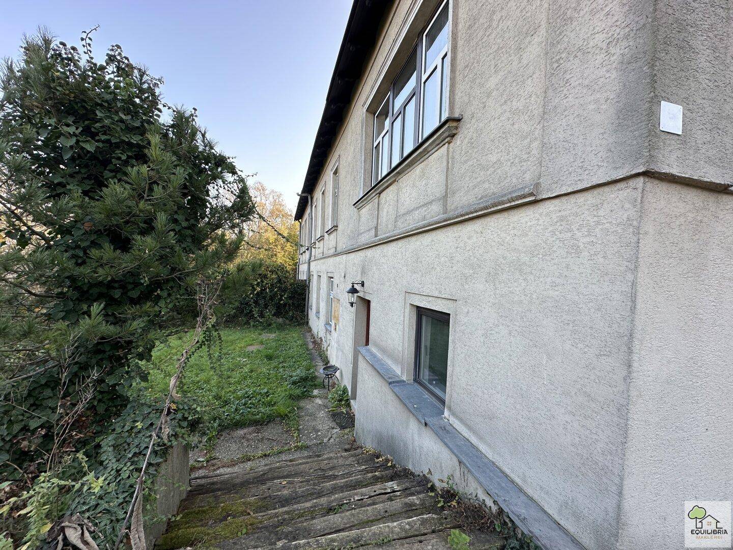 Fassade des Hauses mit Außentreppe, die zum Kellerbereich führt, umgeben von Grün.
